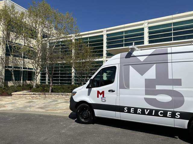 A white service van with the M5 Services logo parked in front of a modern office building with large windows and trees.